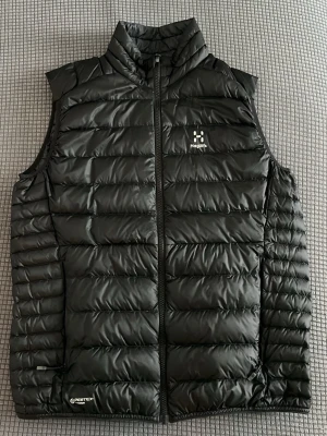 Svart dunväst från Haglöfs - Snygg svart dunväst från Haglöfs med hög krage och dragkedja framtill. Västen har diskret logga på bröstet och är tillverkad i lätt, quiltad design med Pertex-material för extra hållbarhet. Perfekt för lager-på-lager och outdoor-looks.