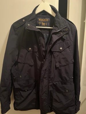 Mörkblå jacka från Woolrich - Snygg mörkblå jacka från Woolrich med fyra stora fickor framtill och dragkedja samt tryckknappar. Jackan har hög krage och axelklaffar, perfekt för dig som gillar en klassisk och funktionell stil. Materialet är slitstarkt och vattenavvisande.