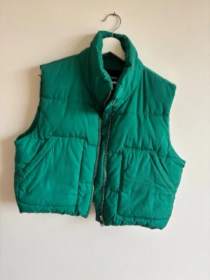 Grön dunväst från Pull&Bear - Snygg grön dunväst från Pull&Bear med hög krage och dragkedja framtill. Västen har två stora fickor på sidorna och en boxig, croppad passform som ger en trendig look. Perfekt att slänga över en hoodie eller tröja för extra värme.