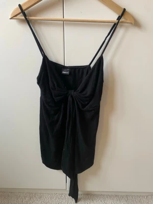 Svart linnetopp med knyt från Gina Tricot - Snygg svart linnetopp från Gina Tricot med tunna axelband och en stor knyt-detalj framtill. Toppen är ärmlös och har en croppad passform som ger en trendig look. Perfekt att styla med jeans eller kjol för en cool outfit.