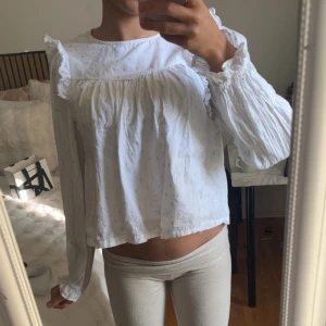 Vit blus med volang från Gina Tricot - Söt vit blus från Gina Tricot med volangdetaljer på axlarna och diskreta guldfärgade prickar. Blusen har lång ärm med resår vid ärmslut och en lös, luftig passform. Perfekt för dig som gillar romantisk och trendig stil.