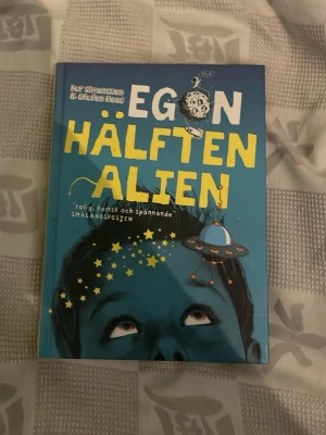 Egon Hälften Alien bok - Rolig och spännande skönlitterär bok med blått och gult omslag, skriven av Åsa Simma och Stefan Roos. Boken har ett färgstarkt tryckt hårdband och illustrationer på framsidan. Perfekt för dig som gillar äventyr och humor.