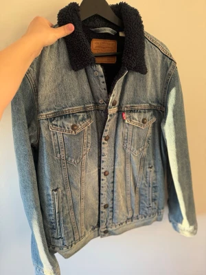 Levi's jeansjacka med teddyfoder - Snygg blå jeansjacka från Levi's med mörkblått teddyfoder och krage. Klassisk modell med två bröstfickor med knapp och två sidofickor. Jackan har knappstängning framtill och detaljer i brunt läder på insidan av nacken.