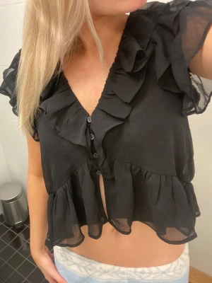 Svart volangblus med knappar - Snygg svart blus med volanger och knappar framtill. Blusen har v-ringning, korta ärmar med volang och är gjord i ett tunt, transparent tyg som ger en luftig känsla. Perfekt att styla med jeans eller kjol för en trendig look.