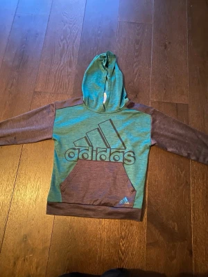 Adidas hoodie storlek M för barn = 10-12år - Snygg hoodie från Adidas i svart och grönt med stor logga på bröstet. Tröjan har huva, magficka och långa ärmar. Perfekt för en avslappnad streetwear-look. Materialet känns mjukt och bekvämt, och designen är sportig med en modern touch. Den ser lite brun ut men det är svart det är bara ljuset som lyser konstigt. 