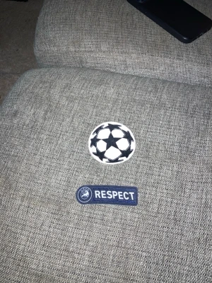 UEFA Champions League tygmärken - Två tygmärken från UEFA Champions League – ett runt med den klassiska stjärnbollen i svart och vitt, och ett rektangulärt med texten 'RESPECT' i vitt på blå botten. Perfekta att stryka eller sy fast på tröjor eller jackor.