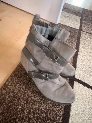 Gråa stövletter med spännen nya! - Snygga gråa stövletter i mockaimitation med dekorativa remmar och metallspännen runt skaftet. Skorna är använda några gånger min lilla syrra tycke först de var bra sen 3 typ dagar efter sa hon de va tajta och ville inte ha de inget är fel med de! Helt rena och inga täcken användning 