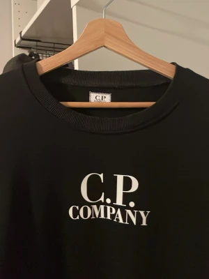 Svart sweatshirt från C.P. Company - Svart sweatshirt från C.P. Company med vit logga tryckt på bröstet. Klassisk rund halsringning och ribbade muddar. Tillverkad i mjukt material som känns skönt mot huden. Perfekt för en avslappnad och stilren look.