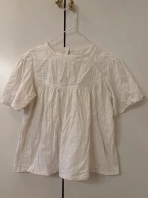 Vit blus med broderad detalj - Söt vit blus med korta ärmar och broderade detaljer upptill. Blusen har en lös passform och rund halsringning samt knapp i nacken. Tillverkad i ett lätt bomullstyg som känns luftigt och bekvämt. Har en ljusgul fläck i armhålan men det syns knappast