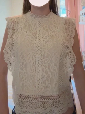 Vit spetsblus med volangdetaljer - Supersöt vit blus i spets med höghalsad krage och vackra volangdetaljer längs ärmhålen och nederkanten. Blusen är ärmlös. Perfekt att styla till jeans eller kjol för en trendig look.