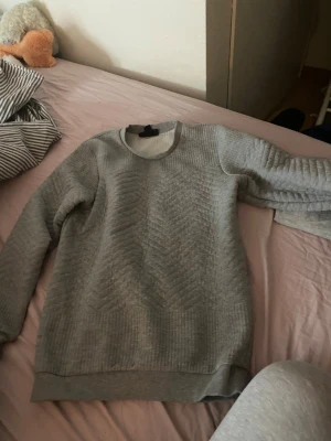 Grå slim fit tröja från SMOG - Säljer en grå slim fit tröja från SMOG i storlek XS. Tröjan har ett diskret mönster framtill och ribbade muddar vid ärmslut och nederkant. Perfekt för en stilren och enkel look.