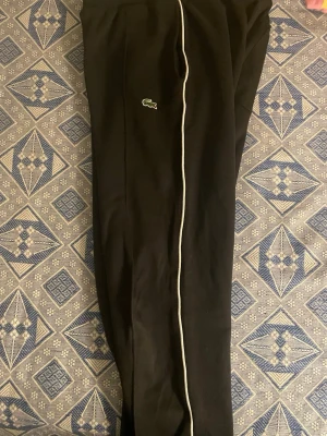 Svarta mjukisbyxor från Lacoste - Säljer ett par svarta mjukisbyxor från Lacoste med regular fit. Byxorna har en vit piping längs benet och den klassiska Lacoste-loggan broderad på sidan. Resår i midjan för skön passform. Perfekta för chill dagar eller när du vill ha en sportig look.