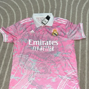 Rosa Real Madrid fotbollströja Adidas - Snygg rosa Real Madrid fotbollströja från Adidas med vit krage och detaljer. Tröjan har ett stort drakmönster i grått över hela plagget och klassiska vita Adidas-ränder på axlarna. Tryck med Emirates Fly Better på bröstet och klubbmärke på vänster sida.
