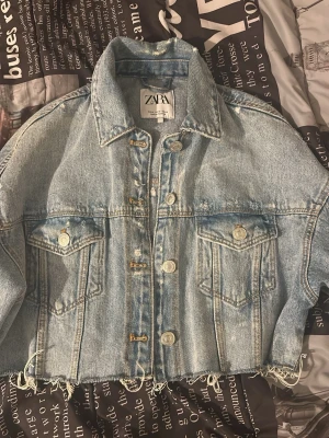 Ljusblå cropped jeansjacka från Zara - Trendig ljusblå jeansjacka från Zara med rå, fransig nederkant och klassisk krage. Jackan har två bröstfickor med lock och silverfärgade knappar framtill. Oversized passform och cool vintagekänsla. Perfekt för dig som gillar streetstyle och denim.