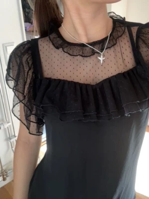 Svart meshblus med volanger - Snygg svart blus med transparent mesh upptill och volangdetaljer över bröstet och axlarna. Prickigt mönster i meshen och rundad halsringning. Perfekt för dig som gillar en trendig och feminin look.