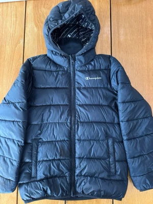 Mörkblå lättdunsjacka Champion barn - Snygg mörkblå lättdunsjacka från Champion med huva. Perfekt för krispiga vårdagar eller som mellanlager. 