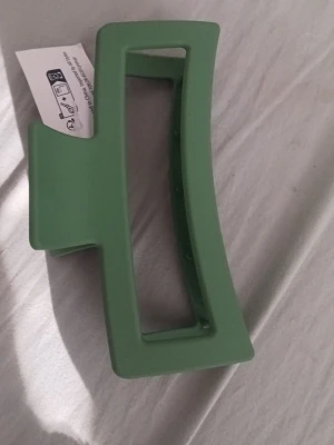 Grön hårklämma i plast - Snygg hårklämma i  grön färg med rektangulär form. Klämman är tillverkad i plast och har breda greppvänliga tänder som håller håret på plats. Perfekt accessoar för att snabbt fixa frisyren. Har prislapp kvar.