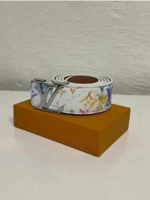 bälte med färgstänk - Säljer ett vitt bälte med färgglada monogramdetaljer i blått, gult och rosa. Bältet har ett stort silverfärgad metall och är gjort i canvas med läderdetaljer. Perfekt accessoar för att lyfta din outfit.