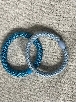 Två flätade hårsnoddar i blå toner - Säljer två runda, tjocka hårsnoddar i flätad design. Ena är ljusblå med vit plastdetalj och den andra är metallicblå med matchande plastdetalj. Båda är gjorda i elastiskt syntetmaterial och passar perfekt för att hålla håret på plats med stil.