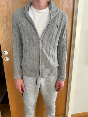 Grå kabelstickad kofta Polo Ralph Lauren - Snygg grå kabelstickad kofta från Polo Ralph Lauren med dragkedja framtill och hög krage. Klassisk logga broderad på bröstet. Stilren helzip tröja som passar till allting! Storlek M. Knappt använt, mest hängt i garderoben då jag växt ur den. Vid fler frågor eller funderingar hör gärna av er!