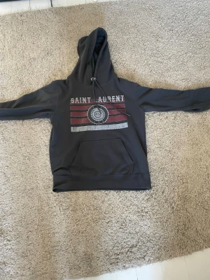 Mörkgrå Saint Laurent hoodie - Snygg mörkgrå hoodie från Saint Laurent med stor huva och dragsko. Framsidan har ett tryck med texten 'SAINT LAURENT' och röda samt vita ränder. Stor magficka framtill. Perfekt för en avslappnad och trendig look.
