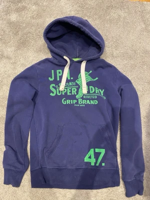 Blå Superdry hoodie med tryck - Mörkblå hoodie från Superdry med grönt tryck framtill och siffran 47 på fickan. Klassisk känguruficka, vita dragsnören och huva. Tillverkad i mjuk bomullsblandning, perfekt för en avslappnad streetwear-look.