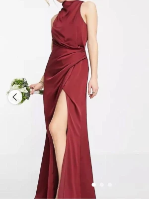 Vinröd klänning med slits - Snygg vinröd långklänning i satinliknande material med hög hals. Klänningen har en draperad detalj i midjan och en slits framtill. Perfekt för bal, bröllop och andra festliga tillfällen. Passar mig som är 165cm och har storlek s/m, använd en gång.