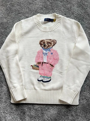 Vit stickad tröja med björnmotiv Polo - Vit stickad tröja från Polo Ralph Lauren med ikoniskt björnmotiv framtill. Björnen har rosa kostym, blå scarf och håller en korg med blommor. Tröjan har ribbade muddar och rund hals. Perfekt för dig som gillar unika och lekfulla plagg.