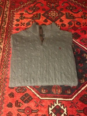 Grå kabelstickad tröja Polo Ralph Lauren - Snygg tröja från Ralph Lauren i en fin grå/grön nyans. Klassisk modell med knappkrage och ikonisk broderad logga på bröstet.  Passar bra både till vardag och lite mer uppklätt – funkar lika bra med jeans som chinos.  Skick: Mycket bra skick, knappt använd.  Detaljer: 	•	Knappkrage och dragkedja 	•	Broderad logga 	•	Mjuk och skön kvalitet