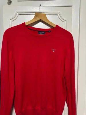Röd stickad tröja från Gant  - Snygg röd stickad tröja från GANT med rund halsringning och diskret logga broderad på bröstet. Tröjan har långa ärmar och ribbade muddar vid ärmslut och nederkant. Perfekt för dig som gillar klassisk och stilren design.