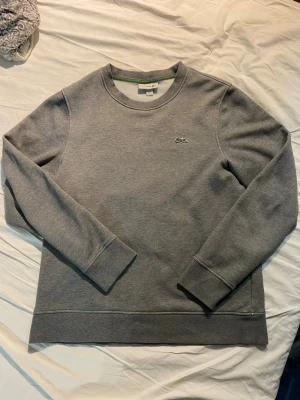Grå sweatshirt från Lacoste - Säljer en stilren grå sweatshirt från Lacoste med det klassiska gröna krokodilmärket broderat på bröstet. Tröjan har rund halsringning, långa ärmar och ribbade muddar vid ärmslut och nederkant. Perfekt för en clean och avslappnad look.