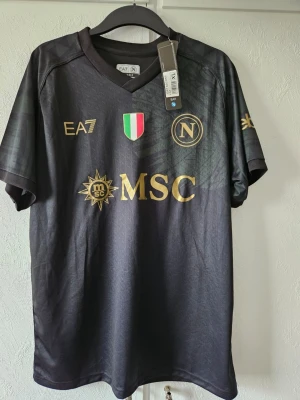 Svart Napoli matchtröja EA7 - Snygg svart matchtröja från Napoli med guldfärgade detaljer och loggor. Tröjan har klubbmärke, italienska flaggan och MSC-sponsor på bröstet. Tillverkad i lätt och ventilerande material, perfekt för fotboll eller träning. (Obs r3pl1ka)