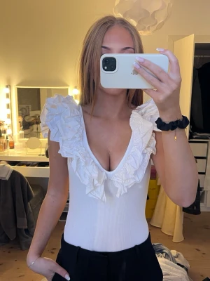 Vit body med volang och v-ringning - Stilren vit body med djup v-ringning och stora volangdetaljer vid axlar och urringning. Tillverkad i ribbat stretchmaterial som sitter tajt och bekvämt mot kroppen. Perfekt för dig som gillar en feminin och trendig look. Bodyn är i storlek L men passar mig som är en S perfekt!