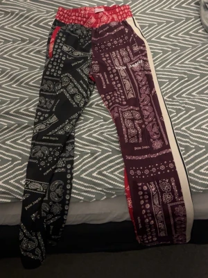 Palm Angels bandana mjukisbyxor - Unika mjukisbyxor från Palm Angels med bandanamönster i svart och vinrött. Ena benet är svart med vita detaljer, andra benet vinrött med vita och röda detaljer samt vit sidoremsa. Resår i midjan och röda fickor. Perfekt för dig som vill sticka ut.