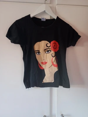 Svart t-shirt med tryck och röd detalj - Cool svart t-shirt med ett stort grafiskt tryck framtill som föreställer en person med en röd blomma i håret. T-shirten har rund hals och korta ärmar. Perfekt för dig som vill sticka ut med en unik design.