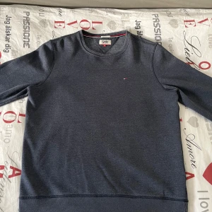 Mörkblå tröja från Tommy Jeans - Säljer en stilren mörkblå tröja från Tommy Jeans i storlek XL. Tröjan har en liten logotyp på bröstet och ribbade muddar vid ärmslut och nederkant. Perfekt för en avslappnad look. Men som man kan se på bilderna är artikeln lite solblek.