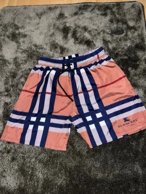 Randiga badbyxor  - Säljer ett par snygga badbyxor från Zara med klassiskt rutigt mönster i marinblått, vitt och rött på en beige botten. De har snörning i midjan och logga på benet. Perfekta för stranden eller poolen!