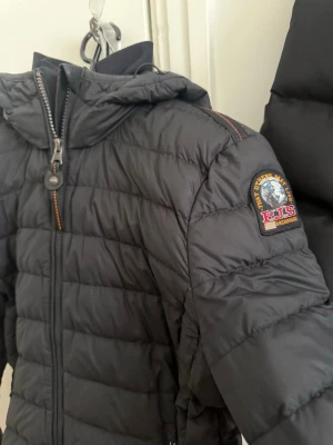 Svart dunjacka från Parajumpers - Snygg svart dunjacka från Parajumpers med huva och dragkedja framtill. Jackan har två sidofickor med dragkedja och det klassiska PJS-märket på ärmen. Lättviktigt material och quiltad design gör den perfekt för kyliga dagar.