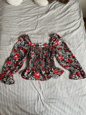 Blommig blus med puffärmar från ONLY - Supersöt blommig blus från ONLY med smockad kropp, volangkant och långa puffärmar. Färgerna är röd, vit, svart och lite grönt. Blusen har fyrkantig ringning och är gjord i ett lätt, vävt material som känns luftigt och skönt.