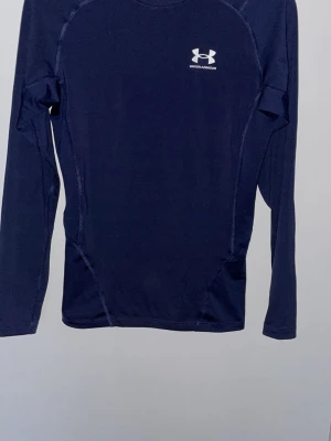 Mörkblå långärmad träningströja UA - Mörkblå långärmad träningströja från Under Armour med vit logga på bröstet. Tillverkad i ett stretchigt funktionsmaterial som andas, perfekt för träning och sportaktiviteter. Tight passform och platta sömmar för extra komfort.