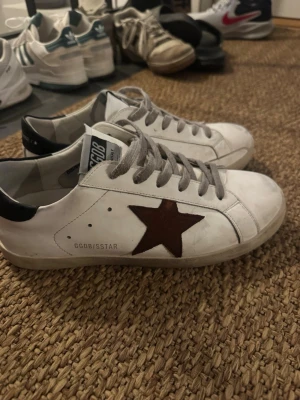Golden Goose vita sneakers med stjärna - Golden Goose sneakers i vitt skinn med brun stjärndetalj på sidan och grå snörning. Klassisk låg modell med rund tå och platt sula. Märkeslogga på plösen och bak på hälen. Snygg och tidlös design som passar till det mesta.