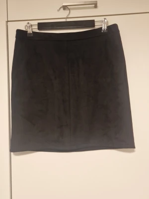 Svart kortkjol i mockaimitation - Stilren svart kortkjol i mjuk mockaimitation. Kjolen har en rak passform och enkel design utan synliga detaljer, perfekt att matcha med olika toppar och jackor. Passar dig som gillar minimalistisk och klassisk stil.