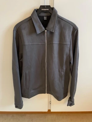 Grå minimalistisk jacka med dragkedja - Stilren grå jacka med rak passform och klassisk krage. Jackan har dragkedja framtill, två sidofickor och knappdetaljer vid ärmsluten. Materialet ser ut att vara mjukt och matt, perfekt för en clean look.