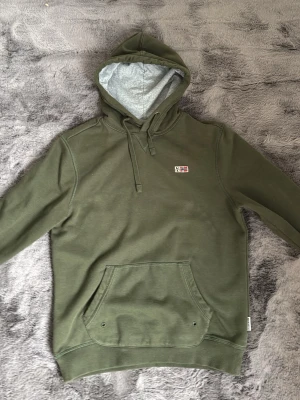  Napapijri hoodie - Snygg olivgrön hoodie från Napapijri med liten logga och norsk flagga på bröstet. Hoodien har en stor känguruficka framtill, dragsko i huvan och grått innerfoder i huvan. Perfekt för en avslappnad och cool stil.