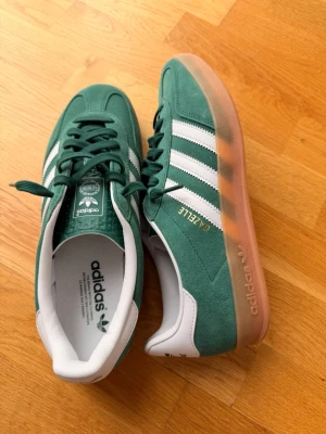 Adidas Gazelle grön/vit sneakers - Snygga Adidas Gazelle sneakers i grön mocka med klassiska vita ränder och detaljer. Låg modell med vit insida och genomskinlig gummisula. Guldfärgad 'GAZELLE'-text. ENDAST ANVÄNDA 2 GÅNGER!!