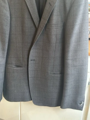 Grå rutig kavaj med diskret mönster - Riktigt Snygg grå kavaj med diskret rutmönster och klassisk passform. Kavajen har två framfickor, bröstficka och två knappar vid ärmslutet. Materialet är vävt och ger en stilren look, perfekt för dig som vill ha en tidlös och trendig stil. 54 slim fit, jag upplever den som liten i storlek, mer 52.