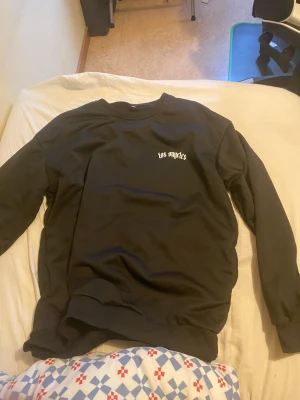 Svart sweatshirt med Los Angeles-tryck - Cool svart sweatshirt med vit text 'los angeles' på bröstet och stort tryck på ryggen. Tröjan har rund halsringning, långa ärmar och ribbade muddar. Perfekt för dig som gillar streetwear och enkel stil.