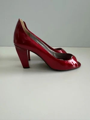 Röda lackpumps med öppen tå - Snygga röda pumps i storlek 39 i glansigt lackmaterial med öppen tå och bred klack. Skorna har en klassisk siluett och ger en riktigt statement-look. Perfekta för dig som vill sticka ut med färg och stil. Innersulan är ljusgrå och klacken är stabil.