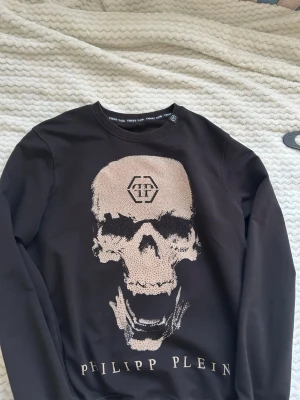 Svart sweatshirt med dödskalle Philipp Plein - Cool svart sweatshirt från Philipp Plein med stor beige dödskalle framtill och logotypdetalj både fram och bak. Rund halsringning och långa ärmar. Perfekt för dig som gillar streetwear och vill sticka ut med en unik design.