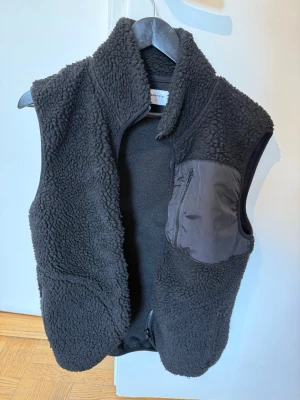 Svart teddyväst Studio Total - Mysig svart teddyväst från Studio Total med hög krage och en praktisk bröstficka i nylon med dragkedja. Västen har en avslappnad passform och är perfekt för lager-på-lager. Materialet är mjuk teddy/fleece och detaljer i nylon ger en cool kontrast.